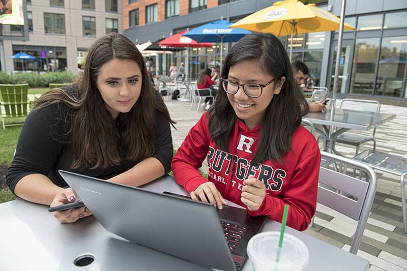 Two students look together at a laptop computer, sitting outside, smiling.