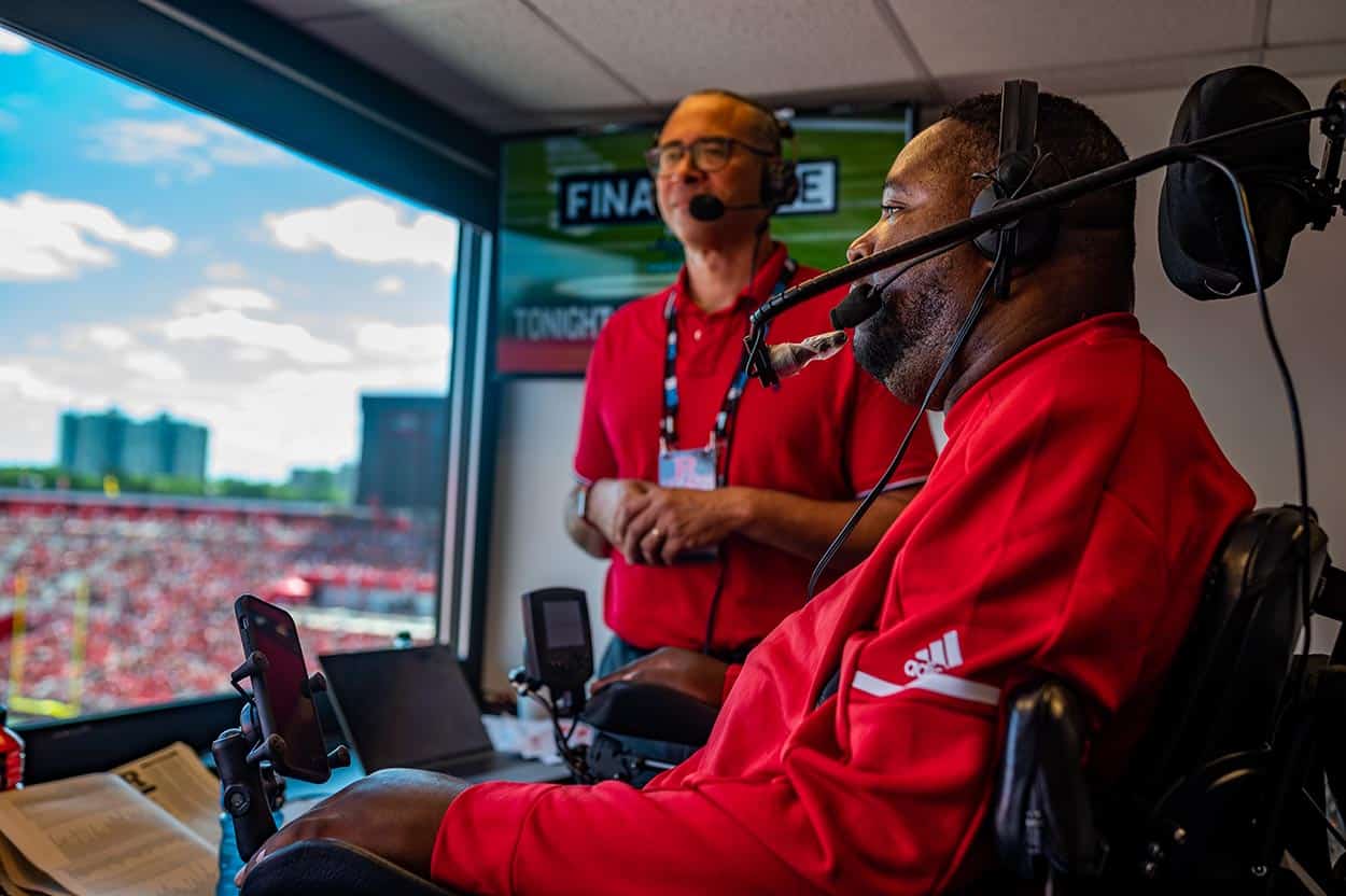 A sports broadcaster sits in a wheelchair at a sporting event, in a booth, with another broadcaster standing beside him.